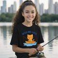 Young girl wearing a black t-shirt with a cat graphic and 'Wish Me Luck' text, holding a fishing rod by a lake with city skyline in the background.