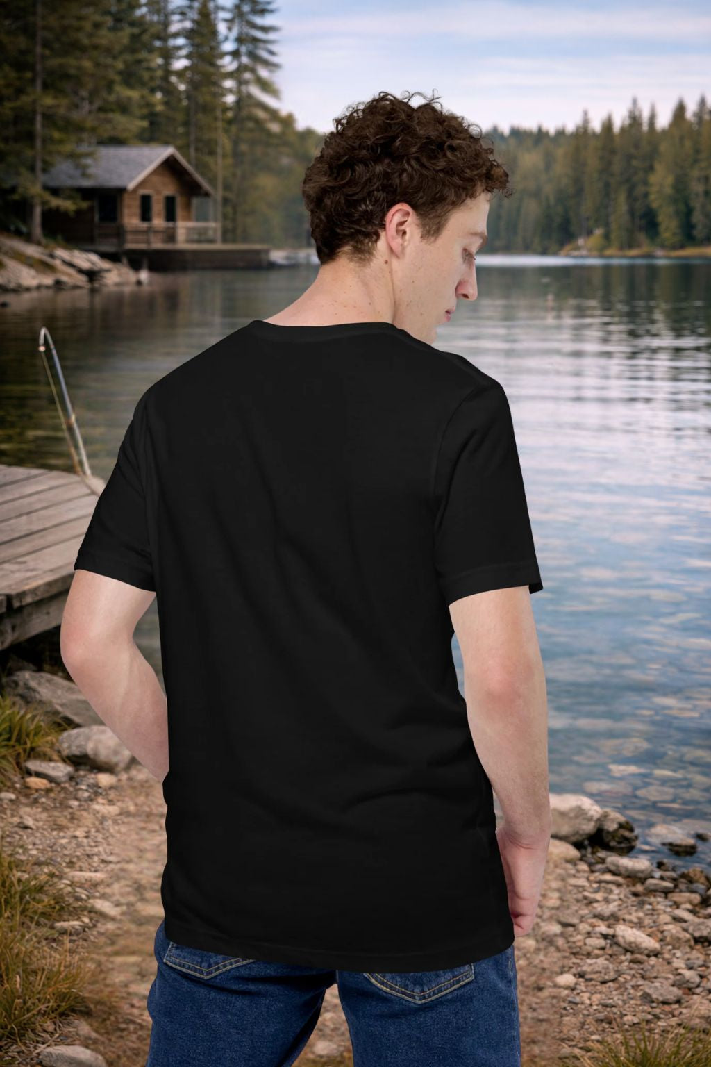 Man wearing a black t-shirt by a lake with a cabin in the background