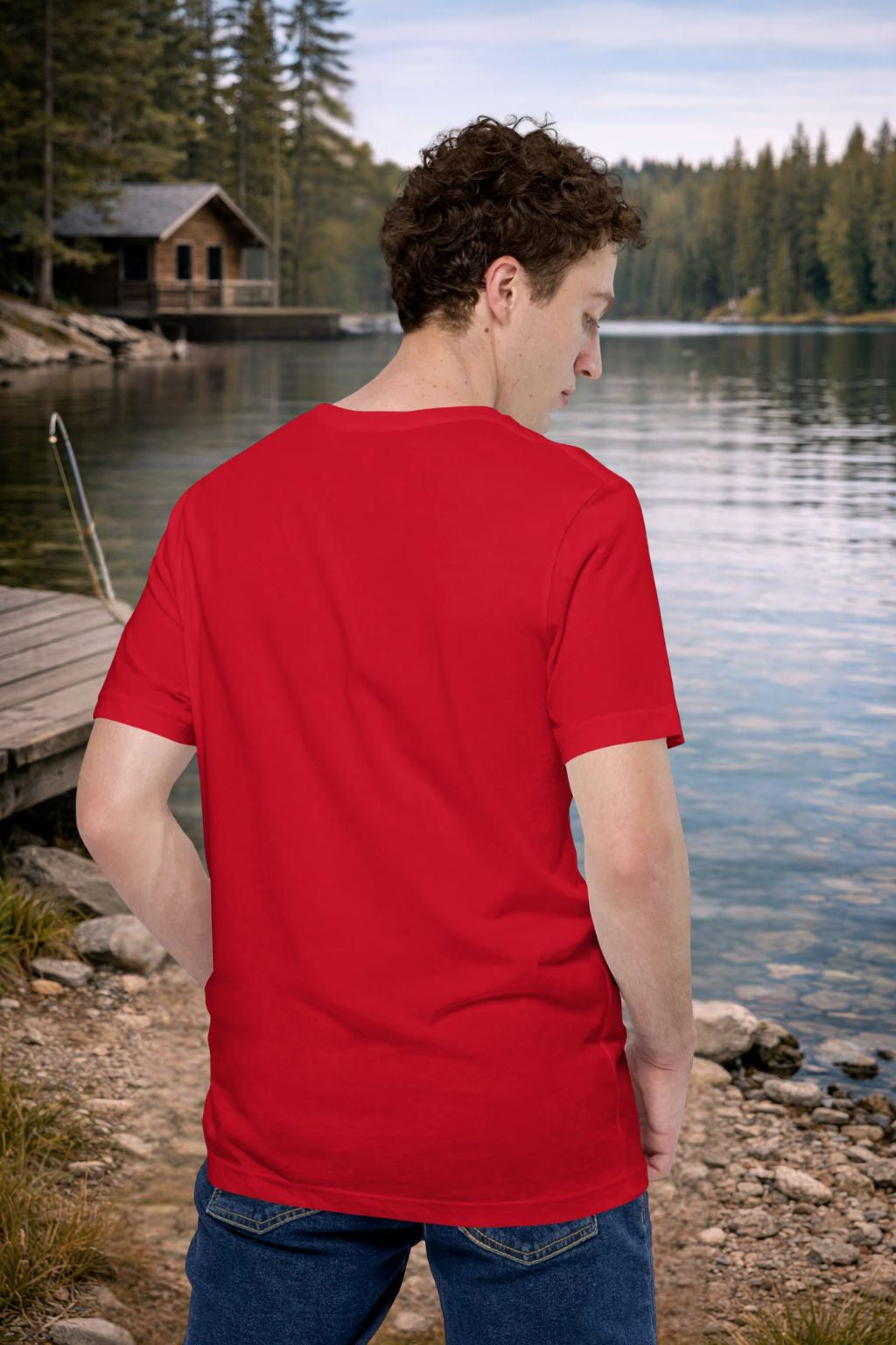 Man wearing a red t-shirt by a lake with a cabin in the background