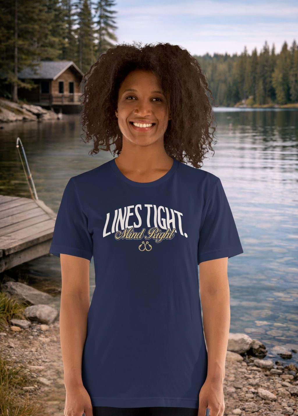 Woman wearing a Fishing navy blue t-shirt with 'LINES Tight' text, standing by a lake with a cabin in the background.