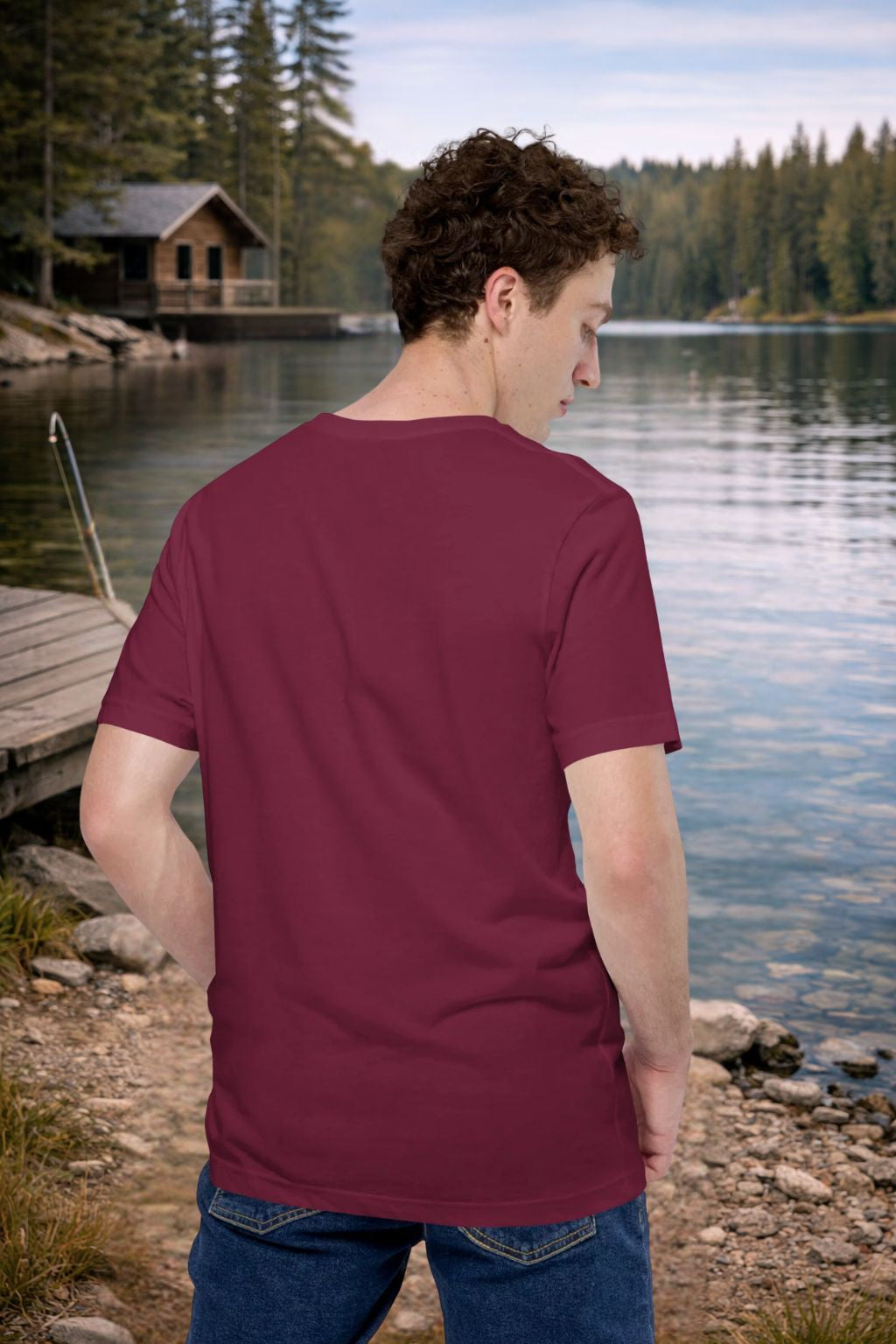 Man wearing a  back maroon t-shirt by a lake with a cabin in the background